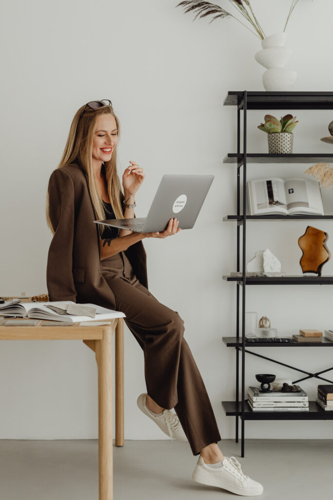 woman in brown suit sitting on desk and holding laptop for video call